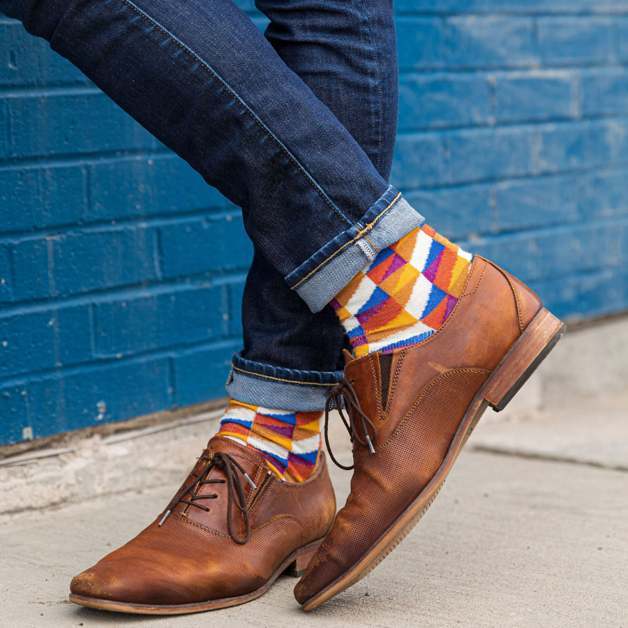 A close up knees-down photo of someone standing in front of a blue brick wall. They are wearing blue jeans, chestnut brown dress shoes, and a pair of Alpacas of Montana white, yellow, gold, orange, purple, and blue triangle pattern casual lounge fashion comfortable soft cozy everyday moisture wicking alpaca wool Swag socks.