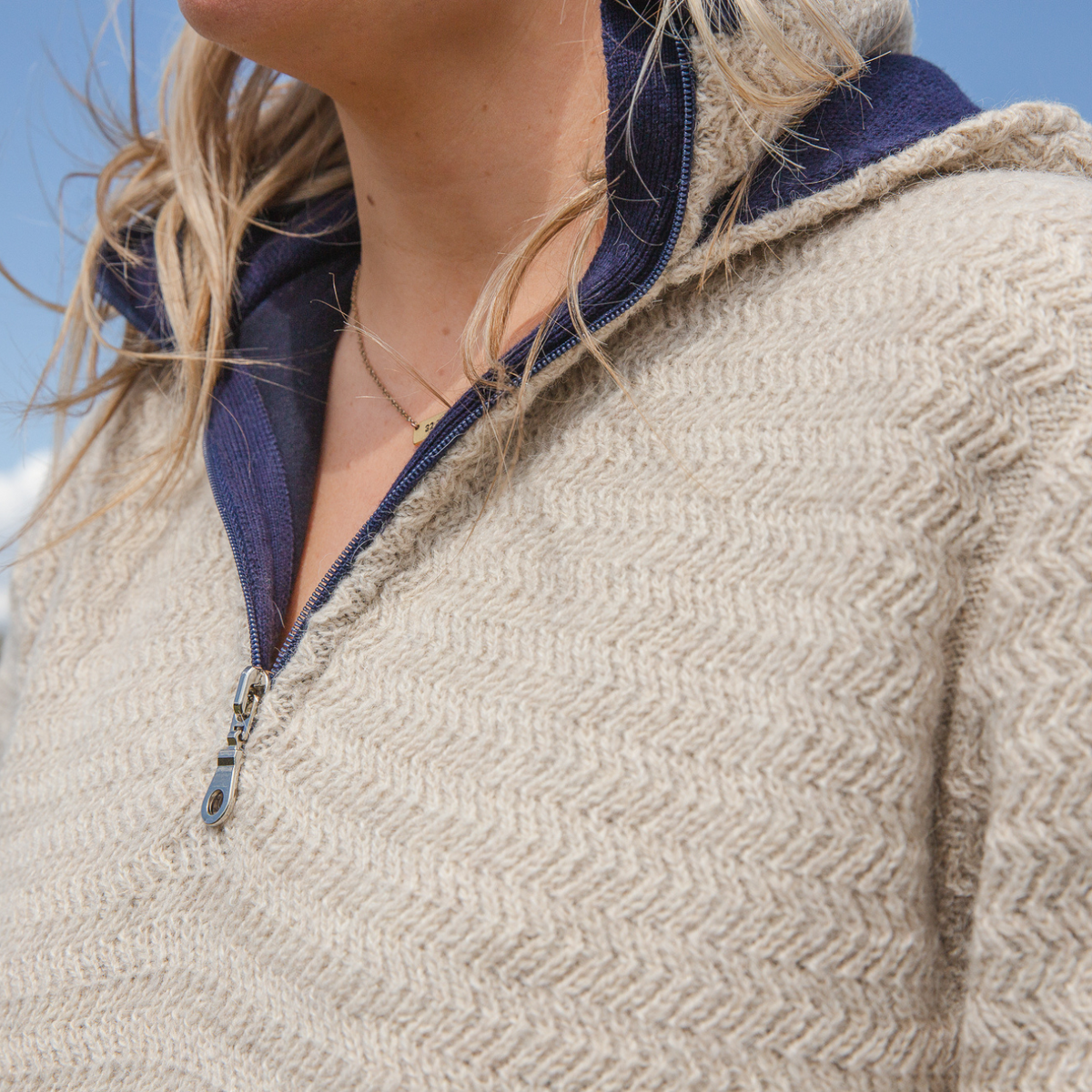 A close up of a blonde woman and the zipper of a sand tan camel oatmeal cream color with navy accents Alpacas of Montana thick warm cozy comfortable soft lined heavyweight warmth moisture wicking antimicrobial stylish women&#39;s fashion oversized alpaca wool hoodie.