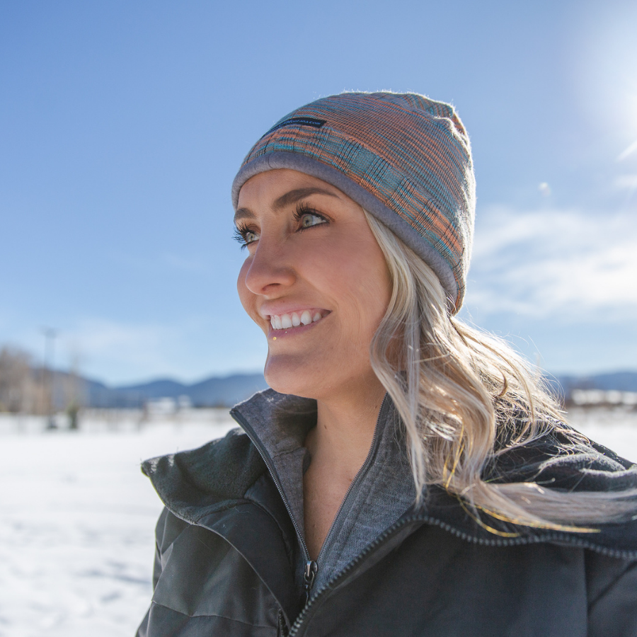 A headshot of a woman standing in a sunny winter scene. She is wearing a graphite color Granite Peak Parka and a teal, orange, and gray cozy soft moisture wicking winter outerwear Backcountry Beanie.