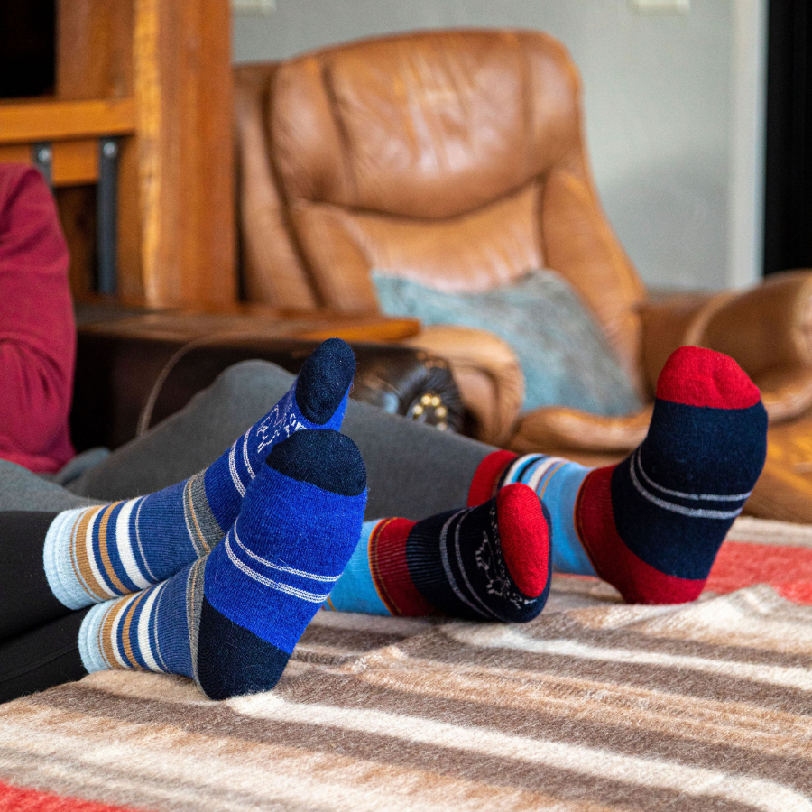 Two pairs of feet of people lounging on a couch wearing soft cozy comfortable moisture wicking lounge and active everyday alpaca wool basecamp socks. The socks on the left are bright blue, navy blue, cobalt, sky blue, gold, and natural white striped. The socks on the right are red, black, sky blue, gold, and natural white striped.