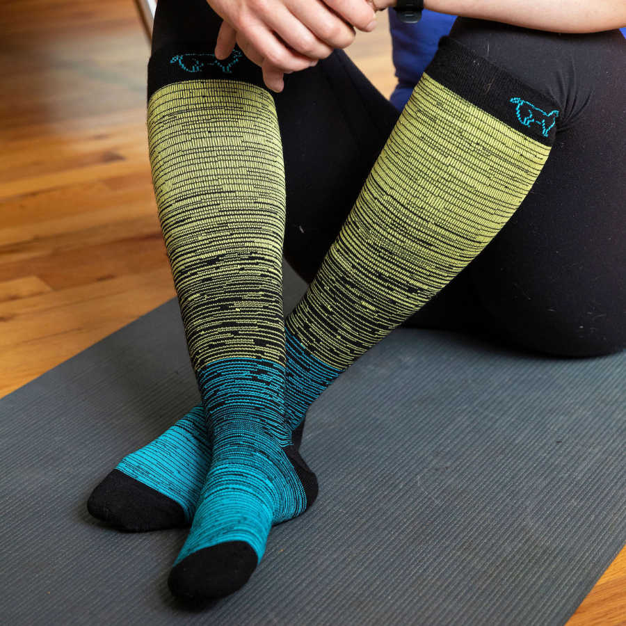 A person sitting on a yoga mat wearing black leggings and the Alpacas of Montana over-the-calf black, yellow, and teal blue compression socks on their crossed feet.