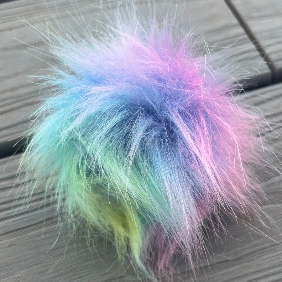 A close up photo of the rainbow tie dye pom pom, a mixture of pastel blue, purple, and green fluff.
