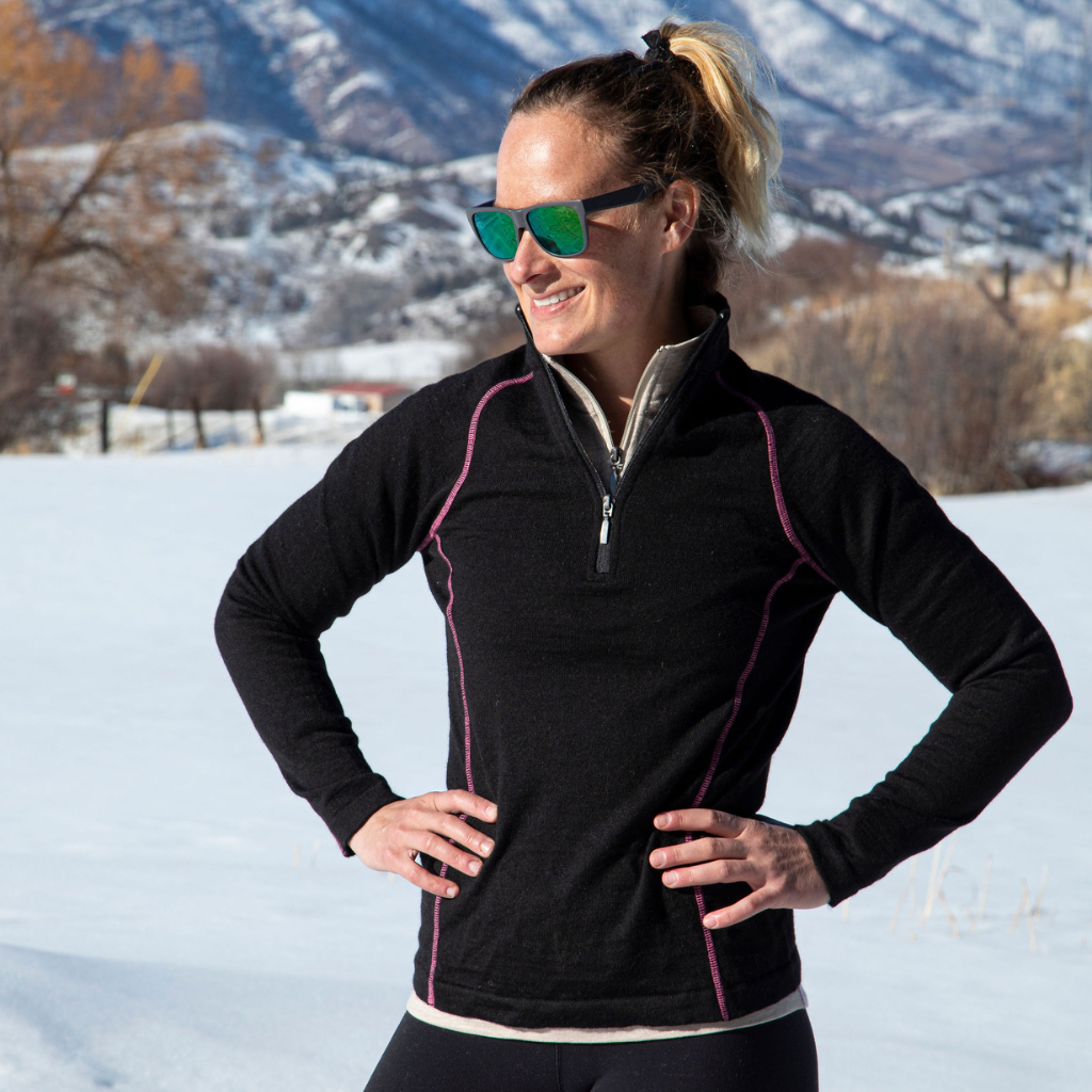 A blonde woman standing with her hands on her hips in a snowy mountain scene. She is wearing sunglasses and a black with pink stitching Alpacas of Montana warm thermal soft cozy comfortable activewear outerwear athletic moisture wicking antimicrobial women's fashion stylish luxury mid-layer quarter-zip alpaca wool long sleeve pullover top for outdoors camping climbing hiking skiing hunting fishing running winter.