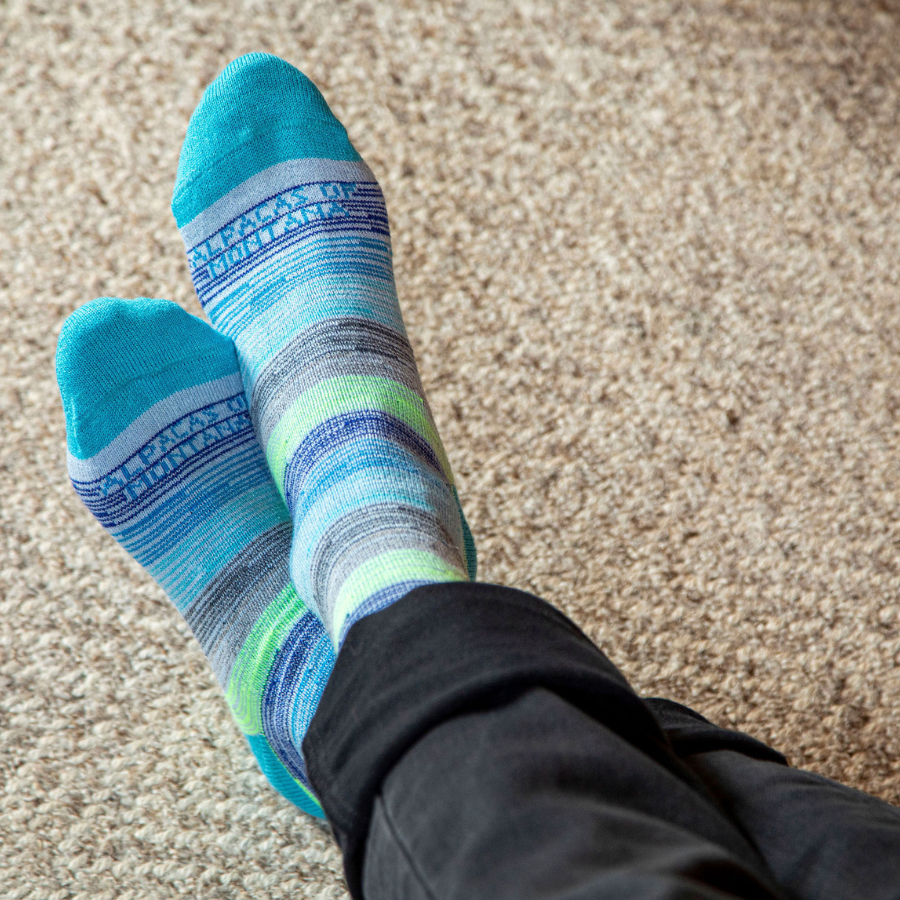 A pair of crossed feet resting on carpet wearing Alpacas of Montana colorful navy, cobalt, sky blue, lime green, gray, and black casual lounge fashion comfortable soft cozy everyday moisture wicking alpaca wool striped socks.