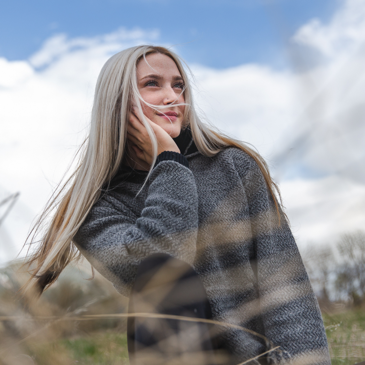 A blonde woman looking into the distance on a sunny day. She is wearing black pants and a medium gray Alpacas of Montana thick warm cozy comfortable soft lined heavyweight warmth moisture wicking antimicrobial stylish women&#39;s fashion oversized alpaca wool hoodie.