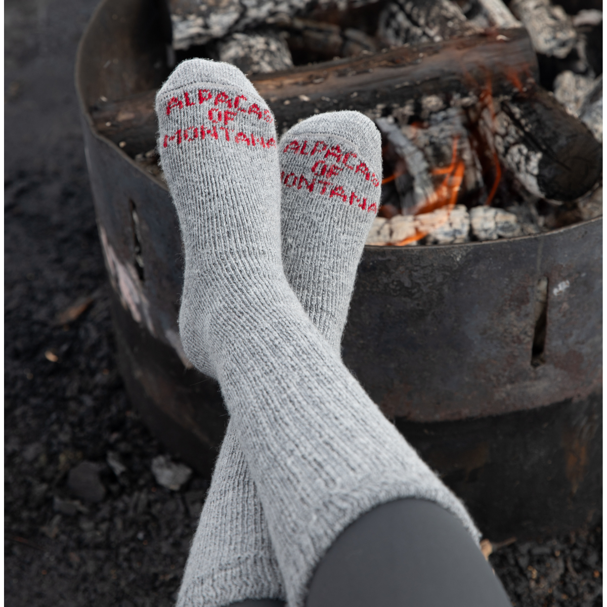 A pair of feet resting up against an outdoor camping fireplace wearing a pair of silver gray Alpacas of Montana cozy comfortable soft warm thermal winter freezing temperatures moisture wicking maximum warmth arctic boot socks for hiking, snowshoeing, hunting, skiing, ice fishing, outdoors.