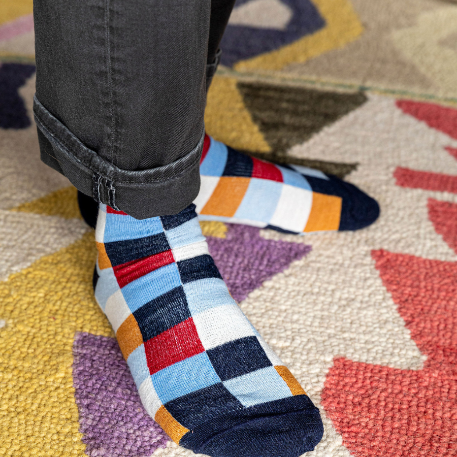 A close up photo of a pair of feet standing on a colorful patterned rug wearing dark gray jeans and a pair of Alpacas of Montana white, red, orange, navy blue, light blue, and sky blue checkered pattern casual lounge fashion comfortable soft cozy everyday moisture wicking alpaca wool Own It socks.