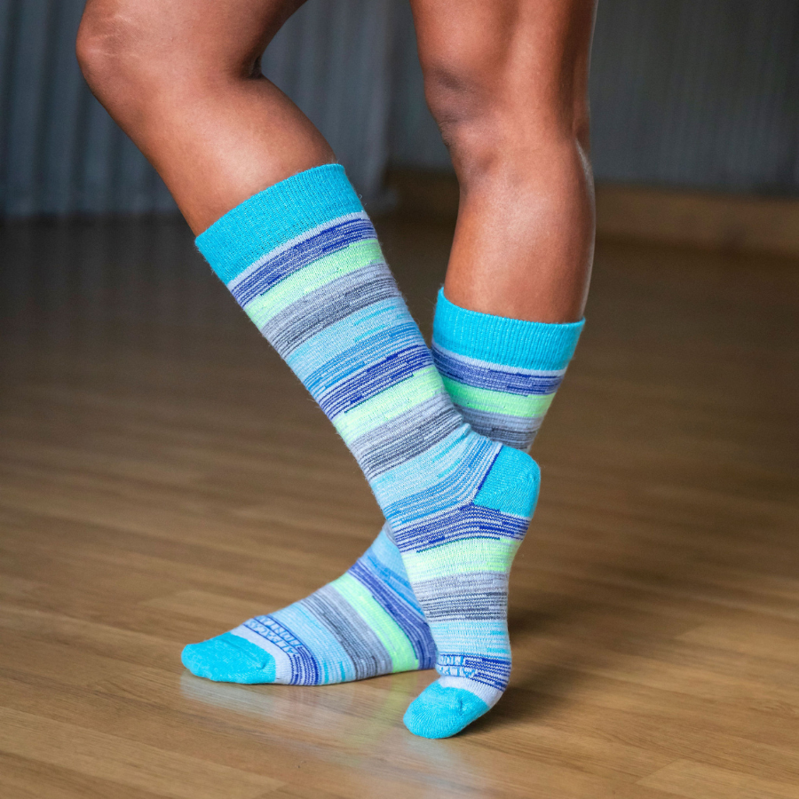 A pair of feet crossed standing on a wooden dancefloor wearing Alpacas of Montana colorful navy, cobalt, sky blue, lime green, gray, and black casual lounge fashion comfortable soft cozy everyday moisture wicking alpaca wool striped socks.