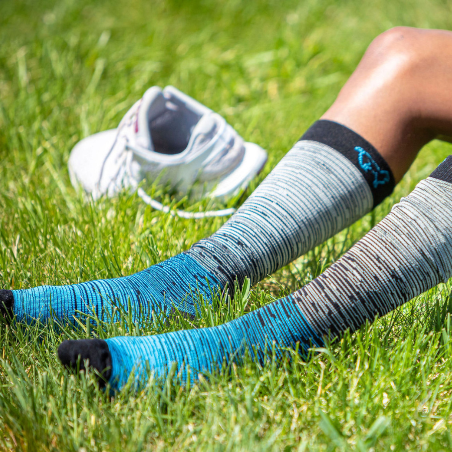 A knees-down photo of a person sitting on grass wearing the Alpacas of Montana over-the-calf black, gray, and teal blue compression socks.