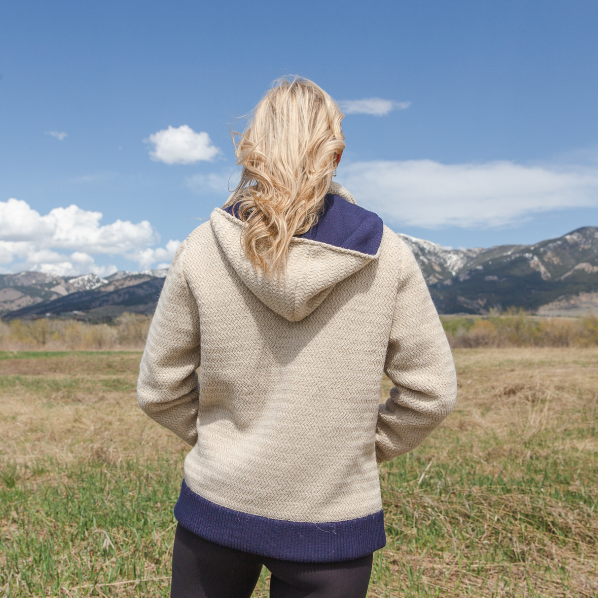 A blonde woman facing away from the camera in front of a sunny mountainous scene. She is wearing black leggings and a sand tan camel oatmeal cream color with navy accents Alpacas of Montana thick warm cozy comfortable soft lined heavyweight warmth moisture wicking antimicrobial stylish women&#39;s fashion oversized alpaca wool hoodie.