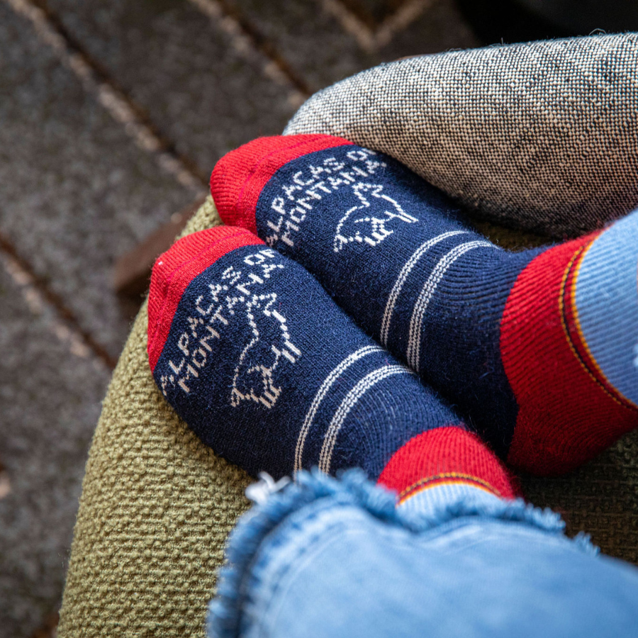 A photo of a person&#39;s feet on a chair wearing blue jeans and a pair of the soft cozy comfortable moisture wicking lounge and active everyday red, black, sky blue, gold, and natural white striped basecamp socks.