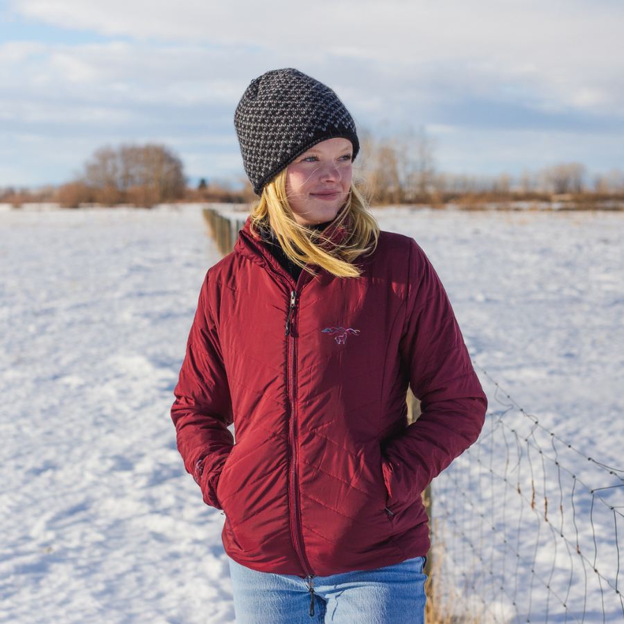 A blonde woman looking to her left in a snowy field. She is wearing blue jeans, a cranberry red Alpacas of Montana women's tempest lite jacket, and a black and gray Alpacas of Montana extremely warm cozy soft windproof comfortable antimicrobial moisture wicking thermal alpaca fleece wool winter fleece lined beanie hat for hiking, skiing, snowshoeing, sledding, outdoors.
