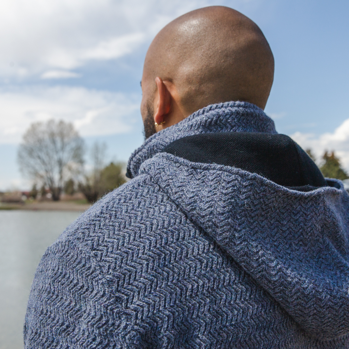 A bald man facing away from the camera on a sunny day. He is wearing a denim navy blue Alpacas of Montana thick warm cozy comfortable soft lined heavyweight warmth moisture wicking antimicrobial stylish men&#39;s fashion oversized alpaca wool hoodie.