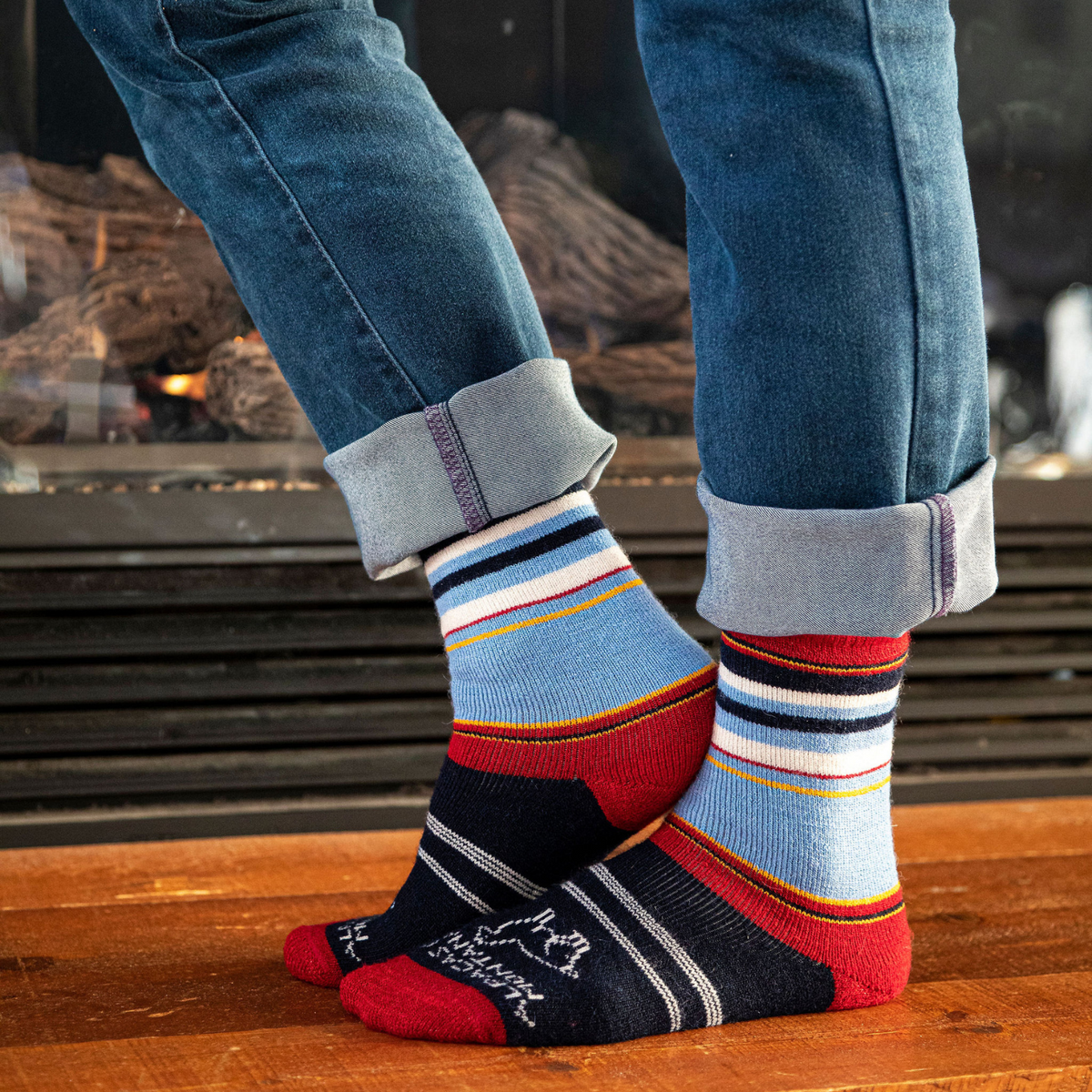 A knees-down photo of a person standing in front of a fireplace wearing blue jeans and a pair of the soft cozy comfortable moisture wicking lounge and active everyday red, black, sky blue, gold, and natural white striped basecamp socks.