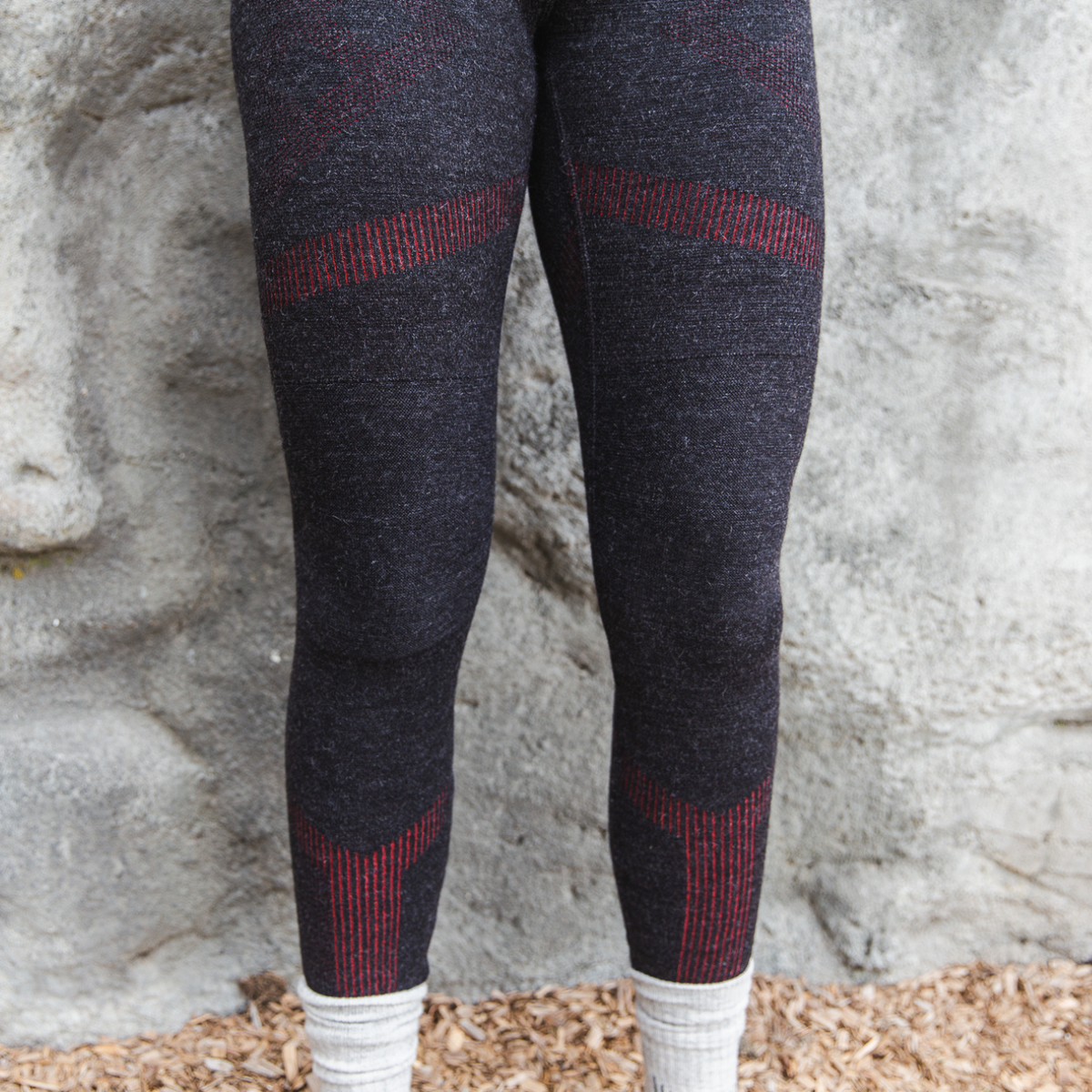 A photo from the thighs down of a woman standing in front of a stone wall wearing a pair of the Alpacas of Montana warm underlayer cozy comfortable thermal moisture wicking long johns winter outerwear heavyweight antimicrobial alpaca wool women&#39;s red and black base layer bottoms for skiing, snowboarding, ice fishing, hunting, camping, arctic, travel