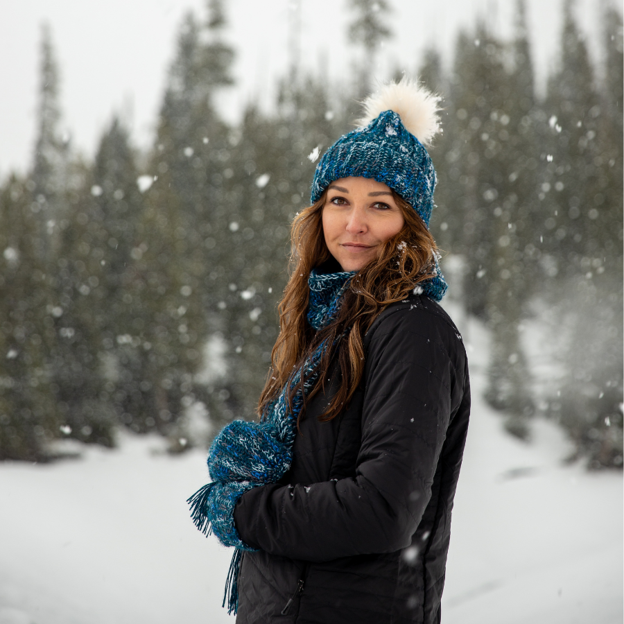 Smiling woman with brown hair standing in winter scene wearing Alpacas of Montana blue knitted crochet handmade matching set of hat, scarf, and ridge ribbed mittens. Handmade items are a mixture of ocean blue, dark turquoise, cobalt, cerulean, gray, and white with long tassels at the end of the scarf and a white pom pom on top of the hat.