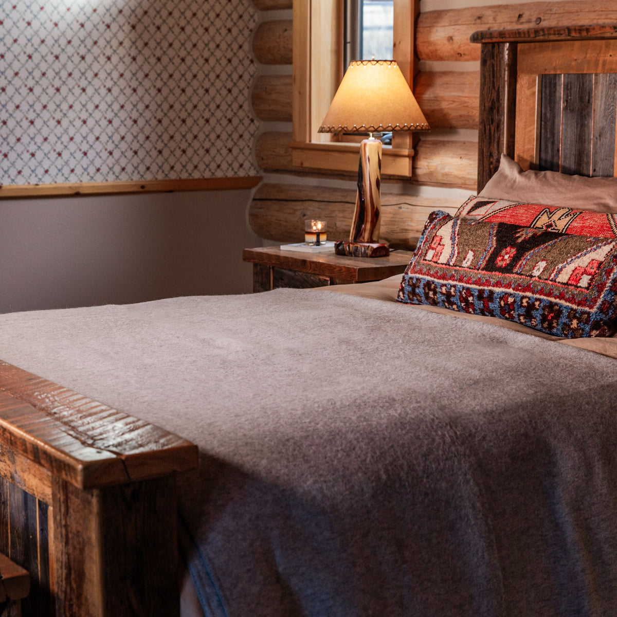A cabin bedroom with a lamp and wooden bedframe. On the bed are several decorative pillows and a mid-gray Alpacas of Montana warm cozy lightweight moisture wicking soft plush king and queen size blanket.