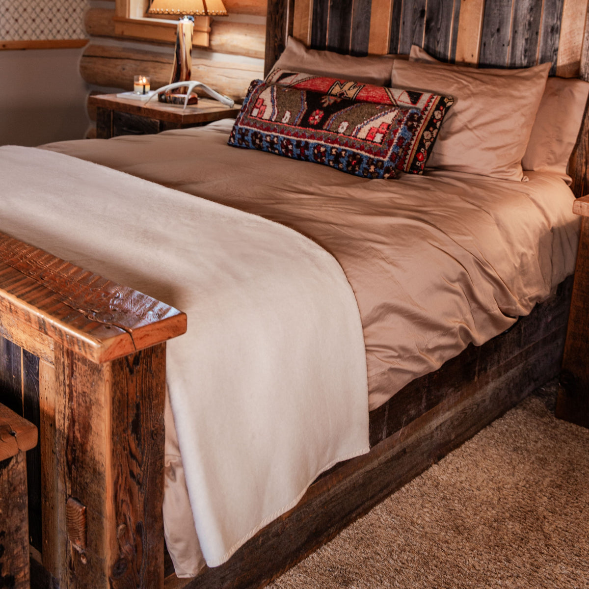 A cabin bedroom with a lamp and wooden bedframe. On the bed are several decorative pillows and a natural white Alpacas of Montana warm cozy lightweight moisture wicking soft plush king and queen size blanket.
