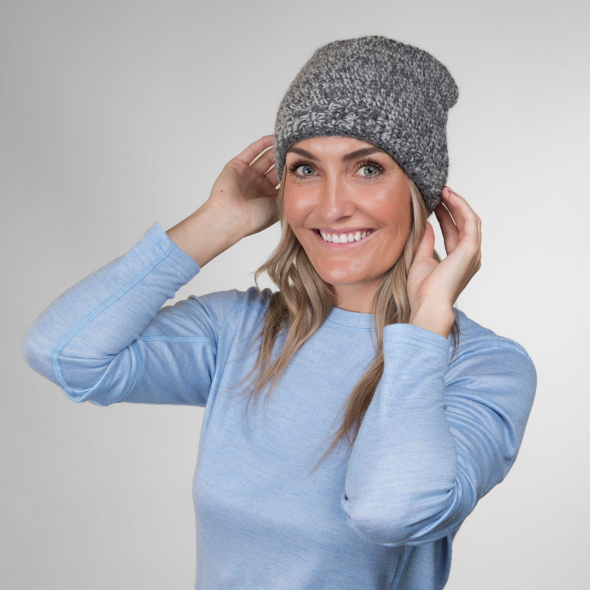 A smiling blonde woman pulling a hat down on her head in front of a white background. She is wearing a light blue long sleeve shirt and an Alpacas of Montana soft cozy comfortable stylish thermal fashionable moisture wicking antimicrobial knitted crochet mountaineer skullcap beanie hat handmade in Montana from light gray and multi-gray alpaca wool and bamboo yarn.