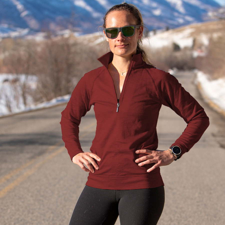 A woman standing with her hands on her hips on a paved road. She is wearing sunglasses, black leggings, and a cranberry cherry red Alpacas of Montana warm soft cozy comfortable activewear outerwear athletic moisture wicking women&#39;s fashion stylish luxury quarter-zip alpaca wool top for outdoors camping hiking skiing hunting fishing running winter.