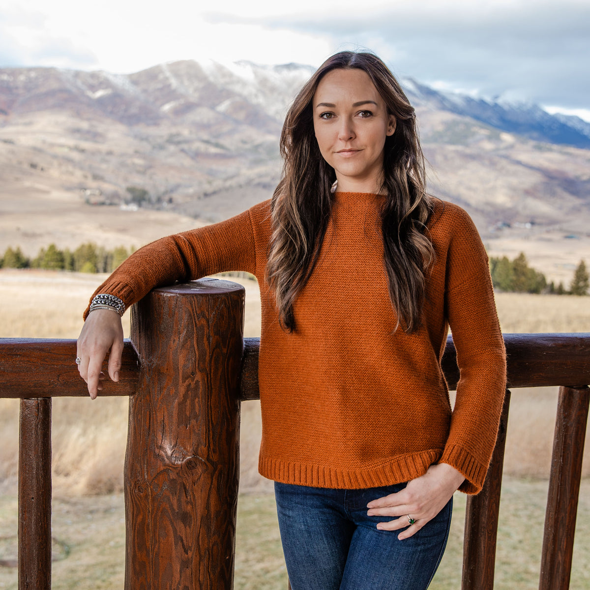 A brown-haired woman leaning against a wooden railing against a mountainous background wearing blue jeans and the Alpacas of Montana soft elegant cute comfortable cozy sweater in the color autumn orange.