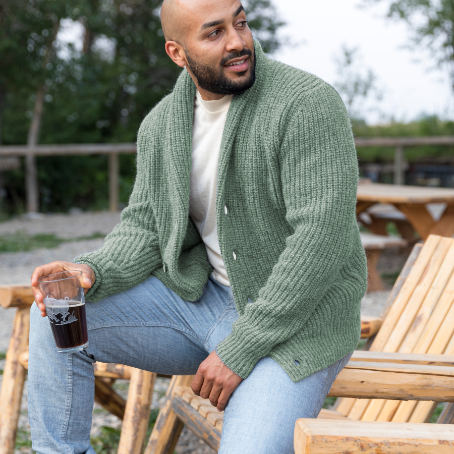 A bald man with a black beard looking off to the side while sitting on a wooden lounge chair holding a glass with a drink in it. He is wearing light blue jeans, a white shirt, and an Alpacas of Montana cozy comfortable soft moisture wicking formal casual stylish upcycled men&#39;s pale mossy green button up chunky knit ribbed cardigan.