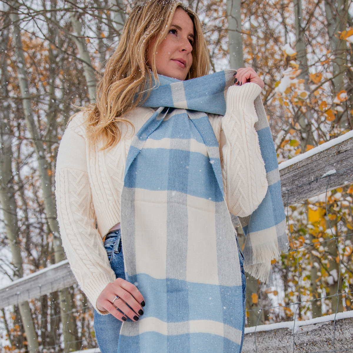 A blonde woman standing next to a wooden fence wearing an ivory woven sweate, blue jeans, and a beautiful soft cozy luxury stylish fashion comfortable accessory sky blue, light gray, and natural white alpaca wool scarf with tassels.