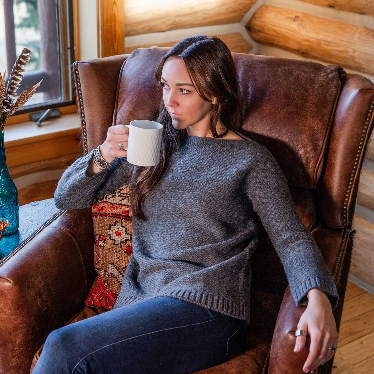 A brown-haired woman sitting in a leather chair inside of a cabin sipping from a white mug wearing blue jeans and a soft elegant comfortable fashionable cozy sweater in the color gray.