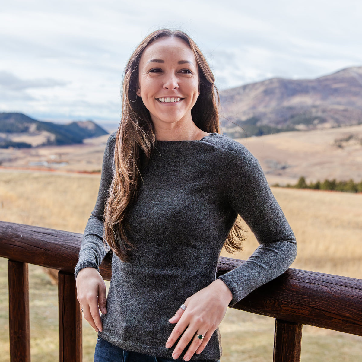 A woman with brown hair leaning against a wooden railing with mountains in the background. She is wearing the comfortable cozy soft elegant fashionable charcoal gray Alpacas of Montana Luxe Light sweater for women.
