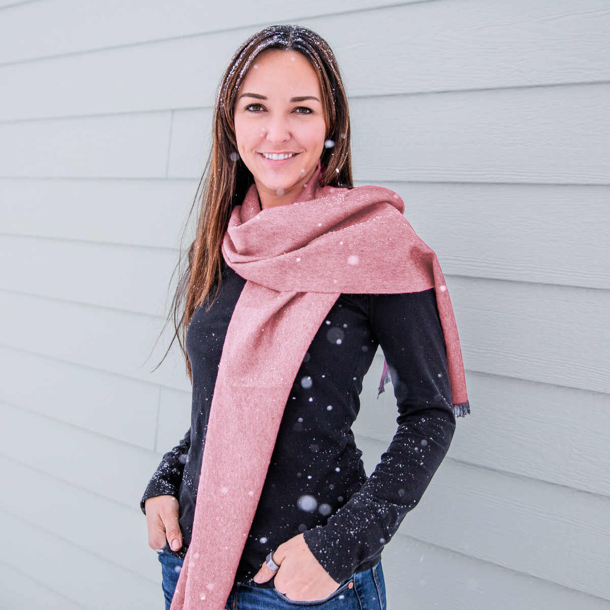 A waist-up photo of a smiling brown haired woman wearing blue jeans, a black long sleeve shirt, and a soft cozy stylish comfortable women&#39;s fashion moisture wicking warm cute lovely herringbone patterned light blush pink and gray alpaca wool scarf.