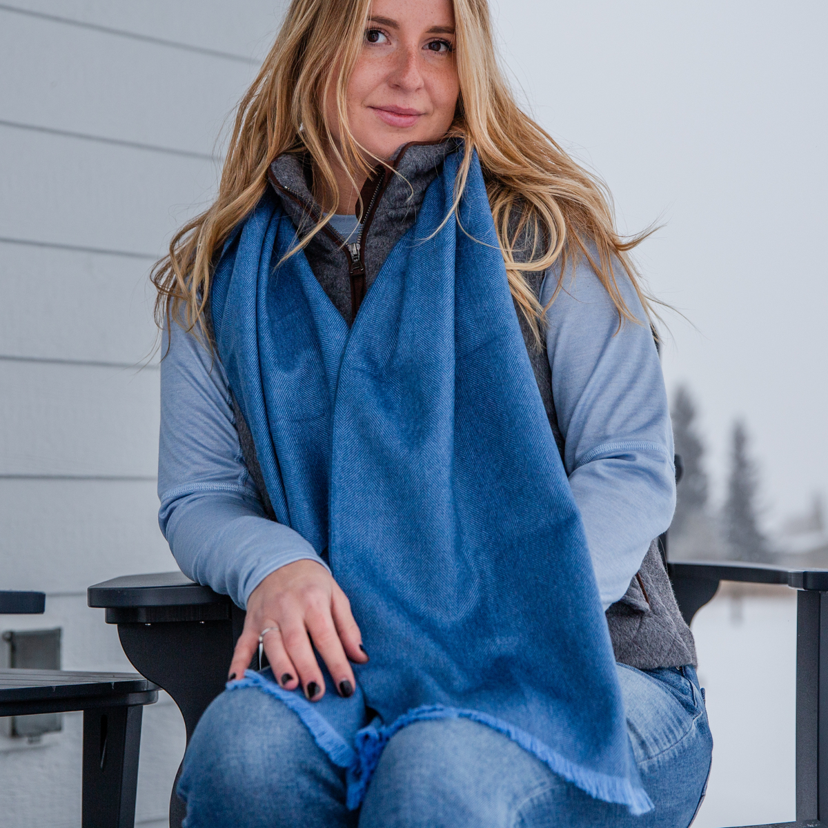 A photo of a blonde woman wearing blue jeans, a light blue long sleeve shirt, a gray Alpacas of Montana hooded vest, and a soft cozy stylish comfortable women&#39;s fashion moisture wicking warm cute lovely herringbone patterned navy blue and sky blue alpaca wool scarf.