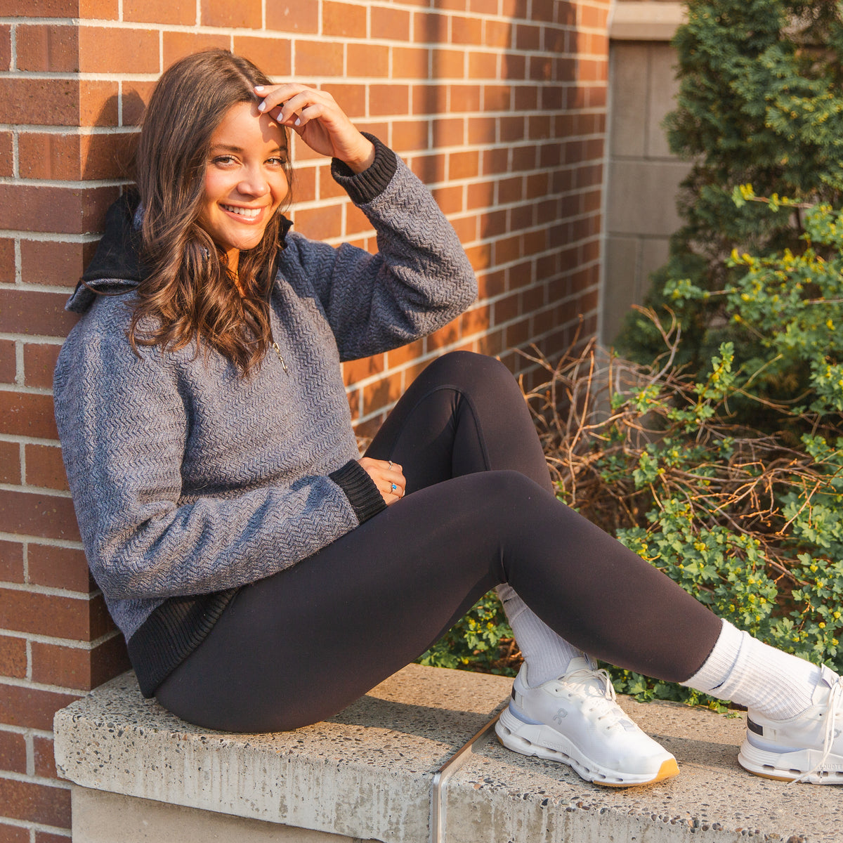 A smiling brown haired woman sitting on a stone wall and leaning against a brick building. She is wearing white socks and sneakers, black leggings, and a denim navy blue Alpacas of Montana thick warm cozy comfortable soft lined heavyweight warmth moisture wicking antimicrobial stylish women&#39;s fashion oversized alpaca wool hoodie.