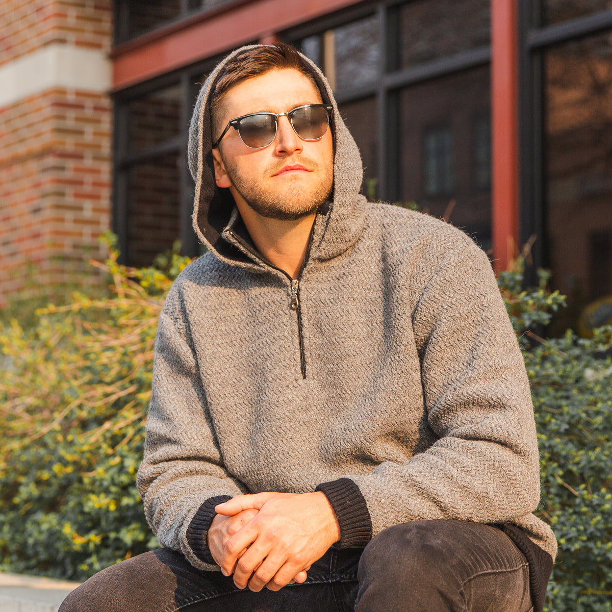 A man sitting in the sun in front of bushes and a building. He is wearing sunglasses, black jeans, and a light gray Alpacas of Montana thick warm cozy comfortable soft lined heavyweight warmth moisture wicking antimicrobial stylish men&#39;s fashion oversized alpaca wool hoodie.