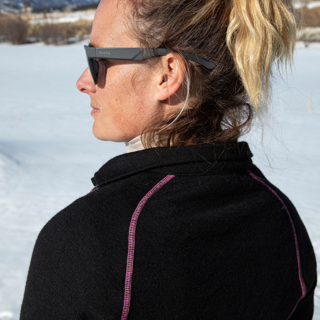 A close up of a blonde woman facing away from the camera in a snowy scene. She is wearing a black with pink stitching Alpacas of Montana warm thermal soft cozy comfortable activewear outerwear athletic moisture wicking antimicrobial women's fashion stylish luxury mid-layer quarter-zip alpaca wool long sleeve pullover top for outdoors camping climbing hiking skiing hunting fishing running winter.