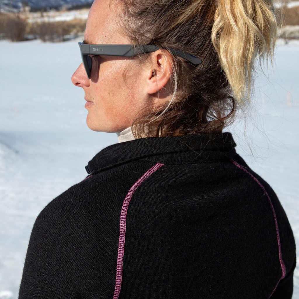 A close up of a blonde woman facing away from the camera in a snowy scene. She is wearing a black with pink stitching Alpacas of Montana warm thermal soft cozy comfortable activewear outerwear athletic moisture wicking antimicrobial women&#39;s fashion stylish luxury mid-layer quarter-zip alpaca wool long sleeve pullover top for outdoors camping climbing hiking skiing hunting fishing running winter.