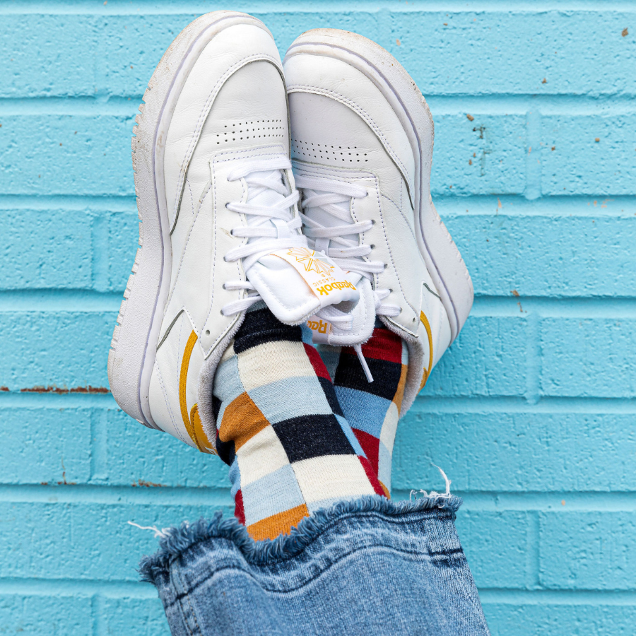 A close up of a pair of feet against a bright blue teal brick wall. The feet are wearing white sneakers, blue jeans, and Alpacas of Montana white, red, orange, navy blue, light blue, and sky blue checkered pattern casual lounge fashion comfortable soft cozy everyday moisture wicking alpaca wool Own It socks.