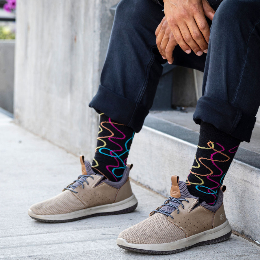A close up photo of a man&#39;s knees and feet sitting on concrete steps. He is wearing tan tennis shoes, black jeans, and the Alpacas of Montana colorful yellow, orange, pink, and blue squiggle pattern black casual lounge fashion comfortable soft cozy everyday moisture wicking alpaca wool Night Life socks.