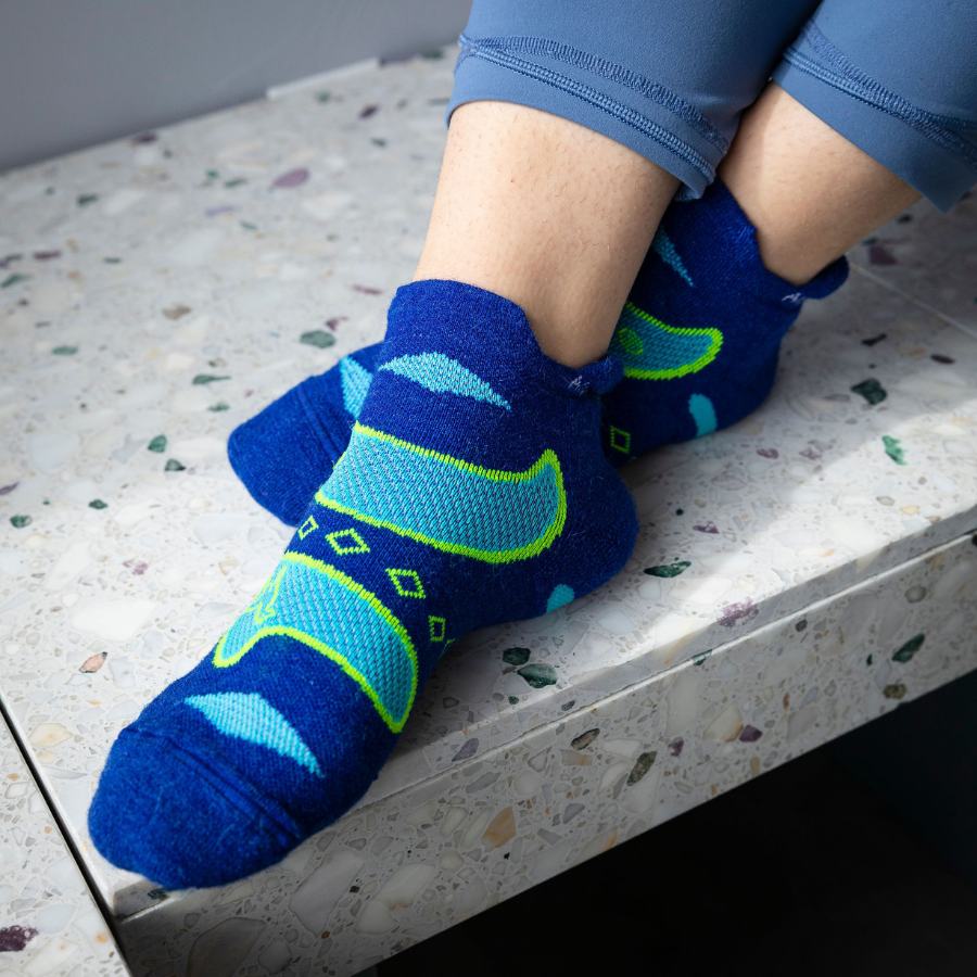 A close up of a pair of feet on a stone bench wearing a pair of Alpacas of Montana soft comfortable breathable athletic outerwear activewear moisture wicking blue, cyan, and lime yellow endurance sock for running, sports, hiking, exercise.