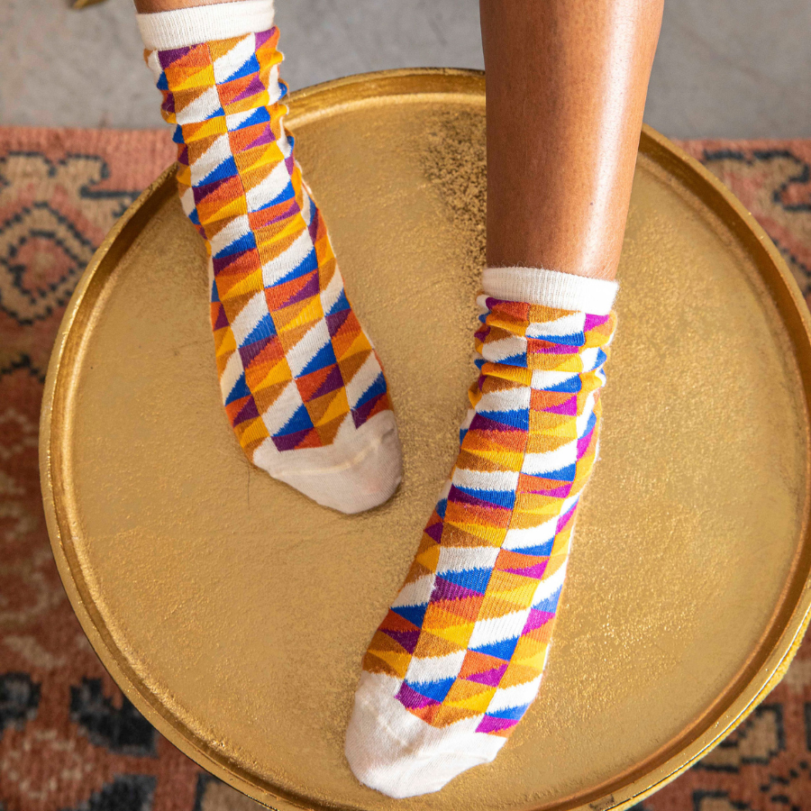 A close up of a pair of feet on a gold stool wearing a pair of Alpacas of Montana white, yellow, gold, orange, purple, and blue triangle pattern casual lounge fashion comfortable soft cozy everyday moisture wicking alpaca wool Swag socks.