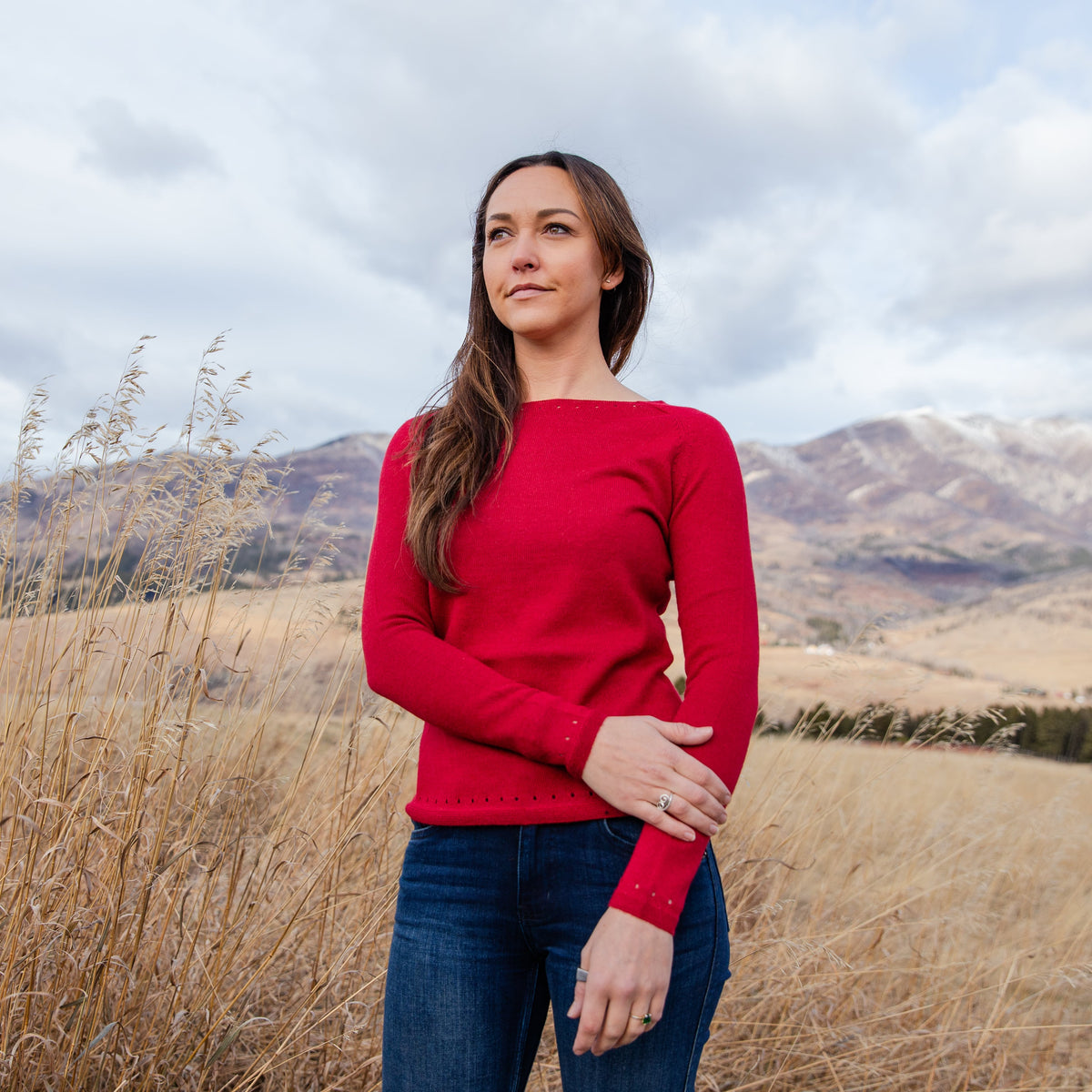 A woman with brown hair stands looking into the distance in a field of golden grass with mountains in the background. She is wearing blue jeans and the comfortable fashionable soft scarlet red Alpacas of Montana Luxe Light sweater for women.