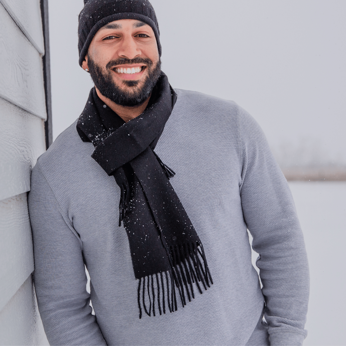 A smiling man with a black beard leaning against a gray house. He is wearing a longsleeve gray shirt, a black Alpacas of Montana backcountry beanie, and an Alpacas of Montana soft stylish women's fashion comfortable cozy cute warm alpaca wool black solid woven scarf with tassels.