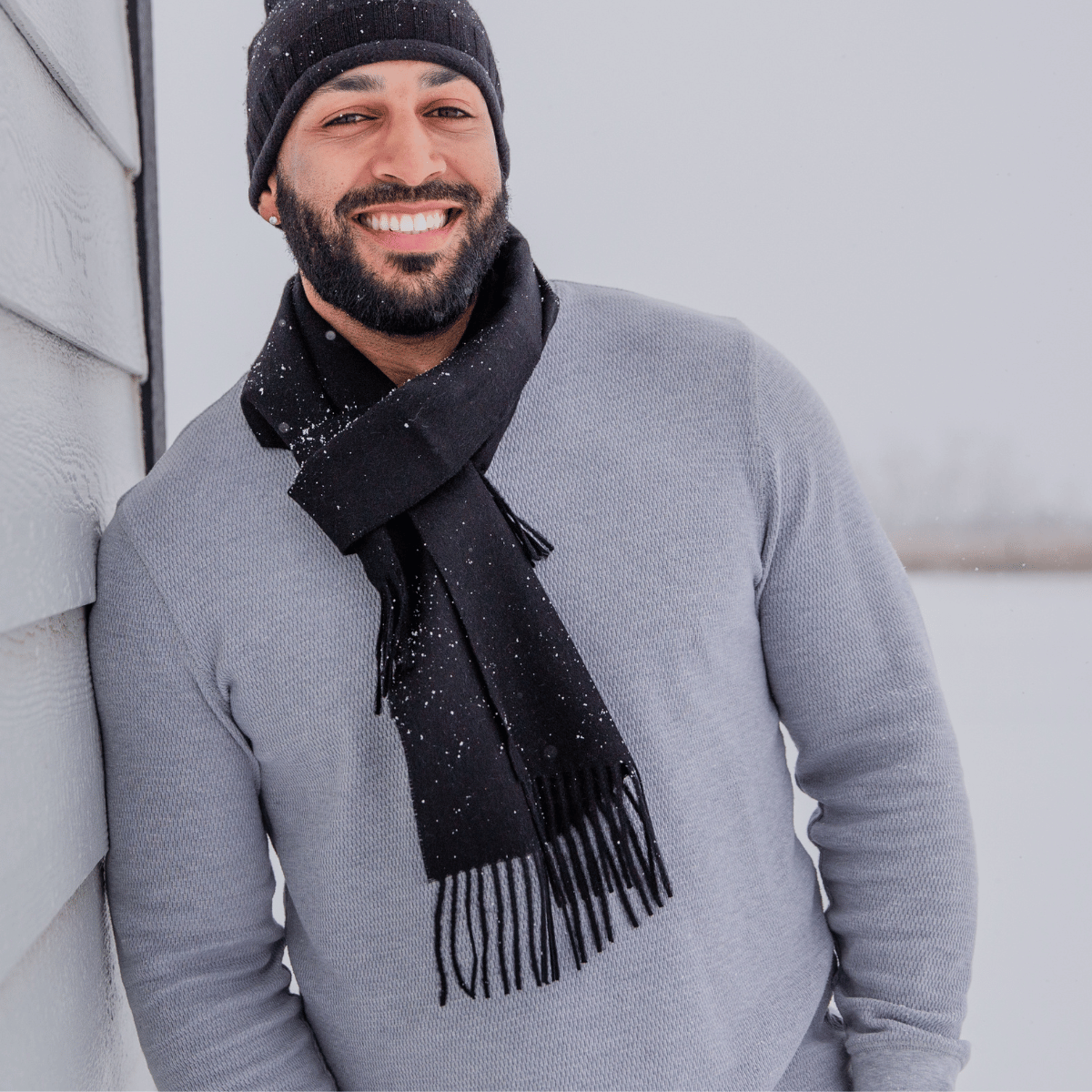 A smiling man with a black beard leaning against a gray house. He is wearing a longsleeve gray shirt, a black Alpacas of Montana backcountry beanie, and an Alpacas of Montana soft stylish women&#39;s fashion comfortable cozy cute warm alpaca wool black solid woven scarf with tassels.
