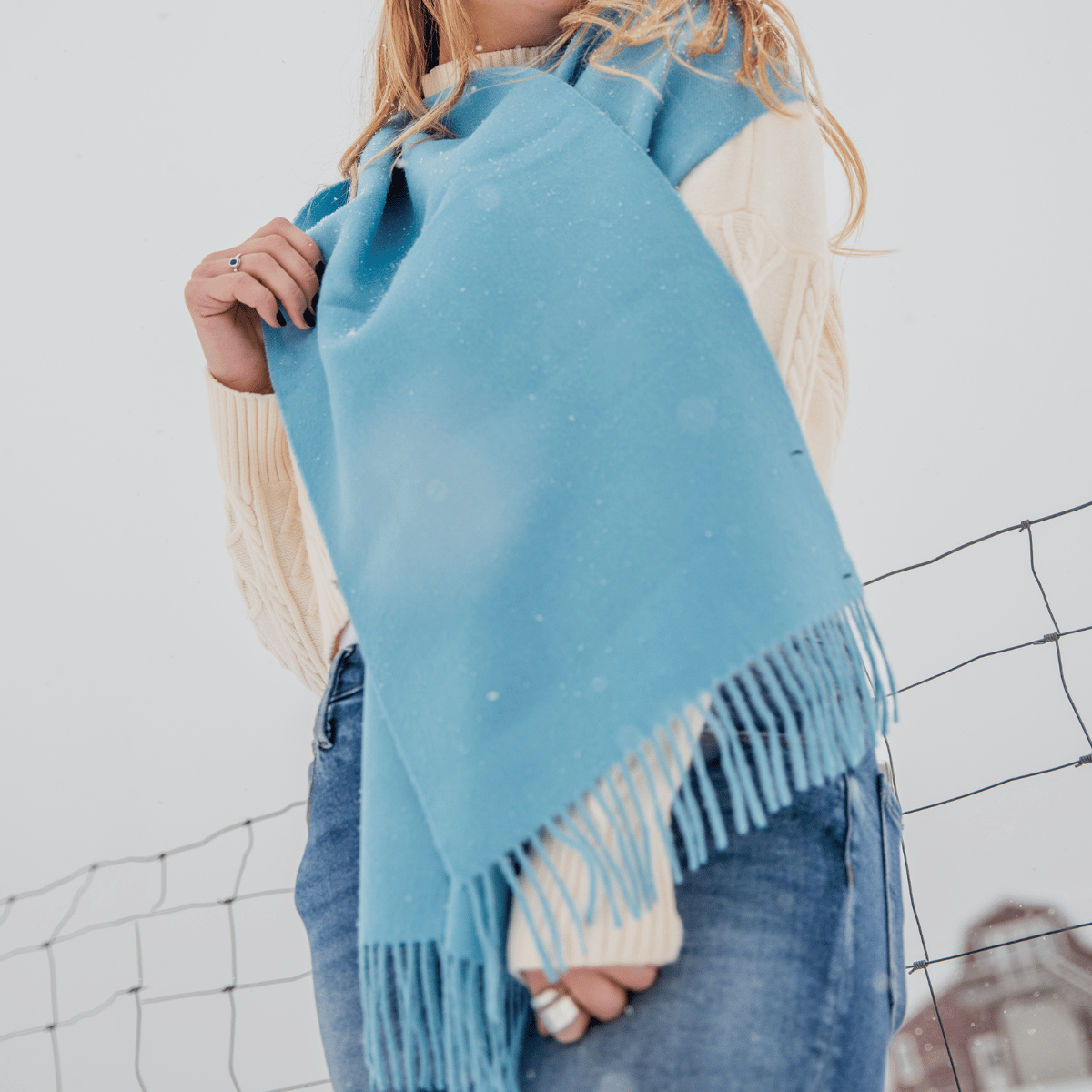 A close up photo of a blonde woman standing in front of a wire fence. She is wearing blue jeans, a cream woven sweater, and an Alpacas of Montana soft stylish women&#39;s fashion comfortable cozy cute warm alpaca wool sky blue cerulean solid woven scarf with tassels.