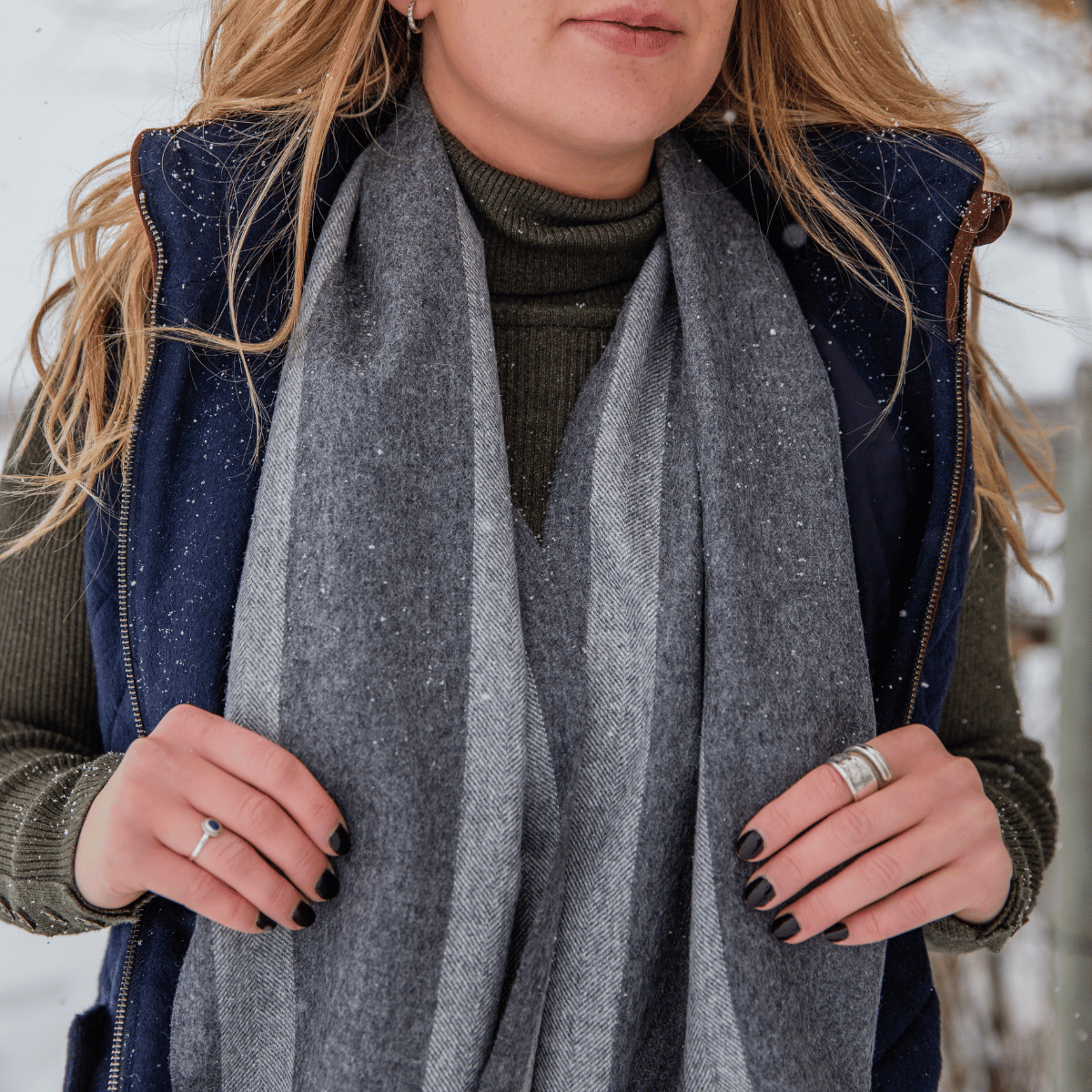 A close up photo of a woman holding the edges of an Alpacas of Montana soft stylish women&#39;s fashion comfortable cozy cute warm alpaca wool light and dark gray stripe solid woven scarf with tassels.