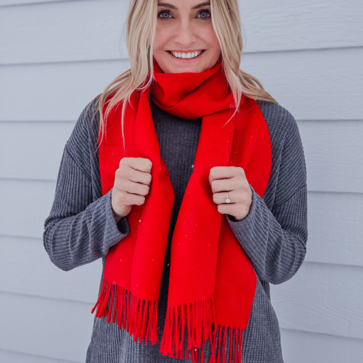 A blonde woman smiling at the camera and wearing a long sleeve dark gray shirt and an Alpacas of Montana soft stylish women&#39;s fashion comfortable cozy cute warm alpaca wool vibrant scarlet red solid woven scarf with tassels.
