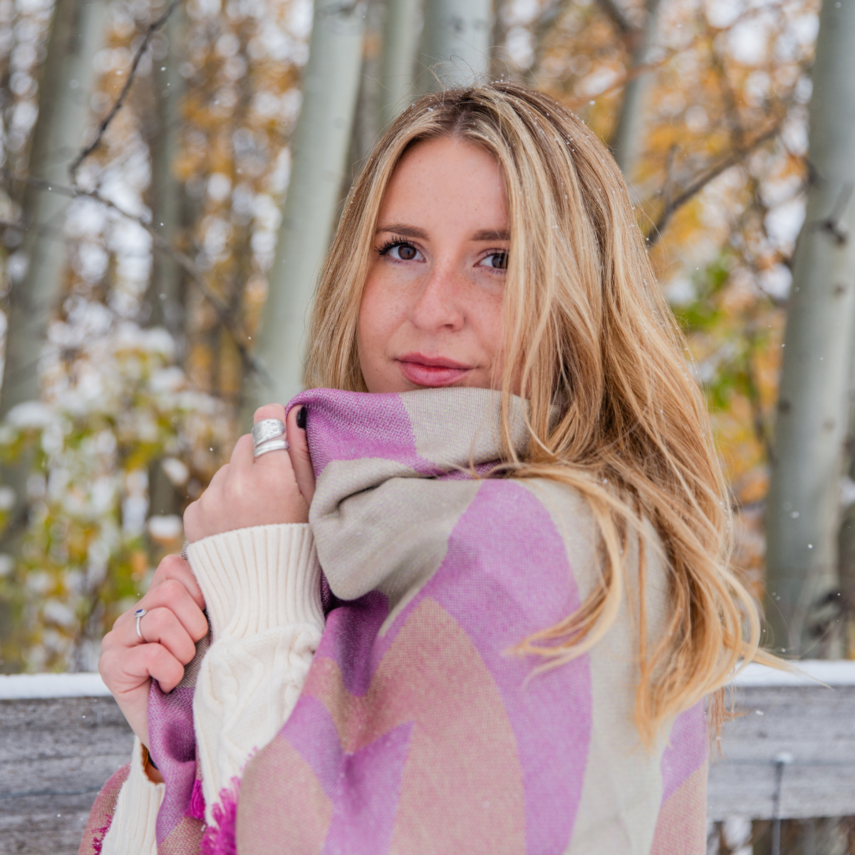 A blonde woman stands in front of a fence and aspen trees with the alpaca and silk soft cozy fashionable pink, green, and orange square shawl wrapped around her shoulders.