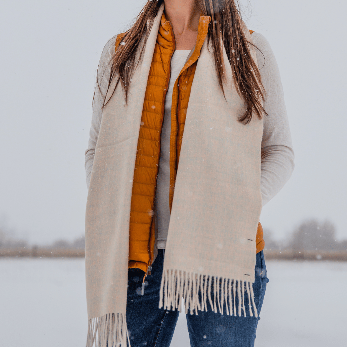 A close up photo of a woman standing in a snowy scene wearing blue jeans, a long sleeve white shirt, an orange vest, and an Alpacas of Montana soft stylish women&#39;s fashion comfortable cozy cute warm alpaca wool taupe color solid woven scarf with tassels.