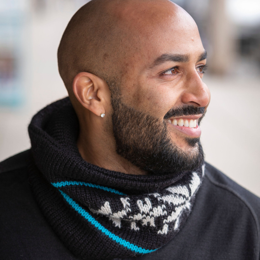 A smiling bald man with a black beard wearing a black shirt and a soft cozy comfortable warm winter thermal moisture wicking heavyweight handmade knit crochet unisex neck gaiter made from natural white, teal blue, and black alpaca wool yarn.