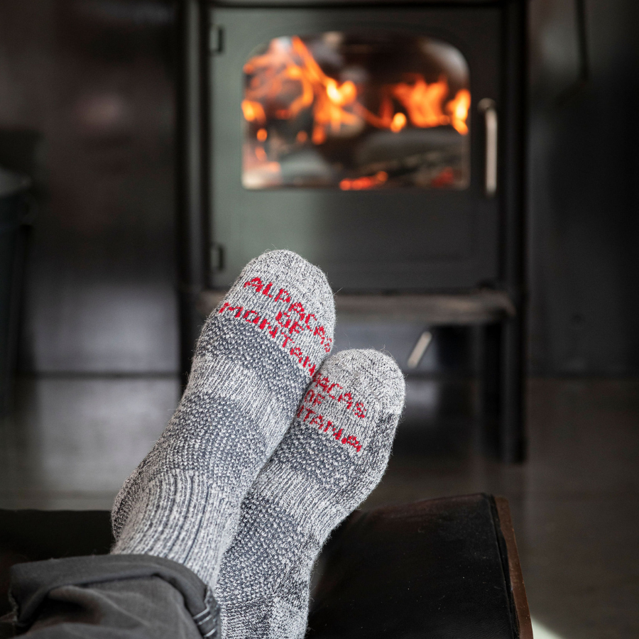 A close up of a pair of crossed feet with a fireplace burning in the background. The feet are wearing Alpacas of Montana cozy soft warm comfortable thermal moisture wicking everyday winter fishing hiking snowshoeing hunting outdoors silver gray extra cushion boot socks.