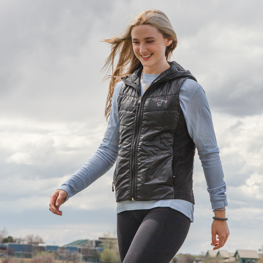 A blonde woman walking in gray leggings, a light blue long sleeve shirt, and a black Alpacas of Montana lightweight athletic activewear outerwear breathable moisture wicking antimicrobial soft comfortable exercise alpaca women&#39;s stretch lite vest for running sports exercise work out hiking climbing camping biking